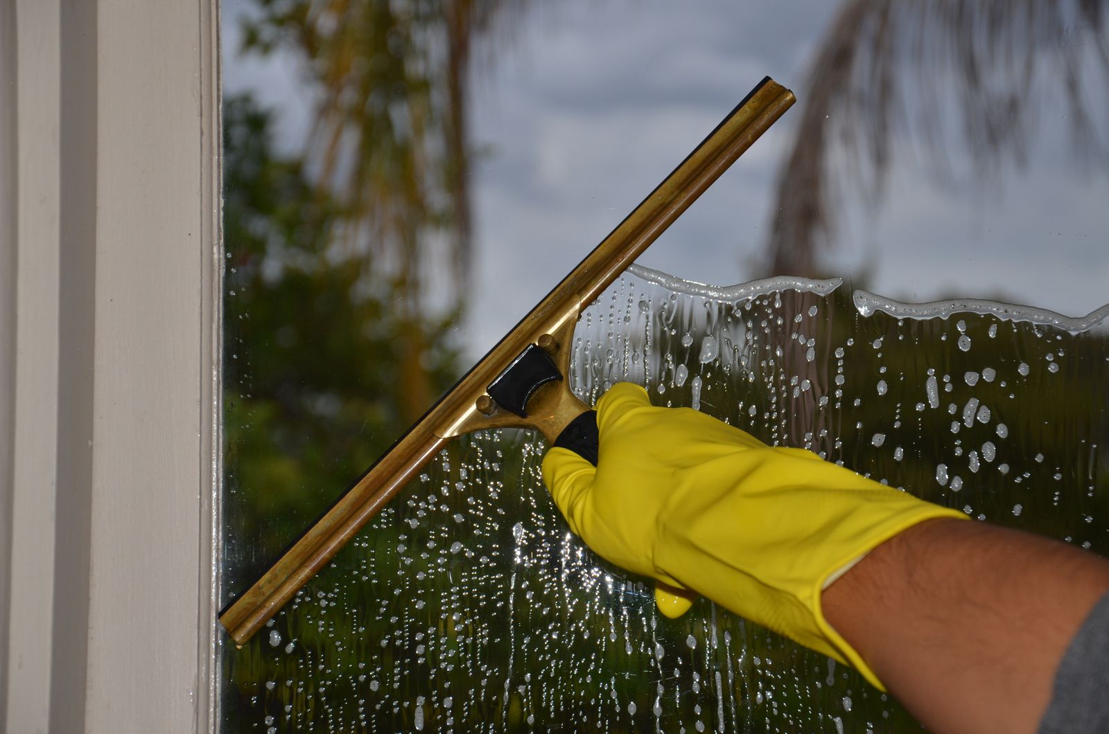 Close-up of a gloved hand cleaning a soapy window with a brass squeegee